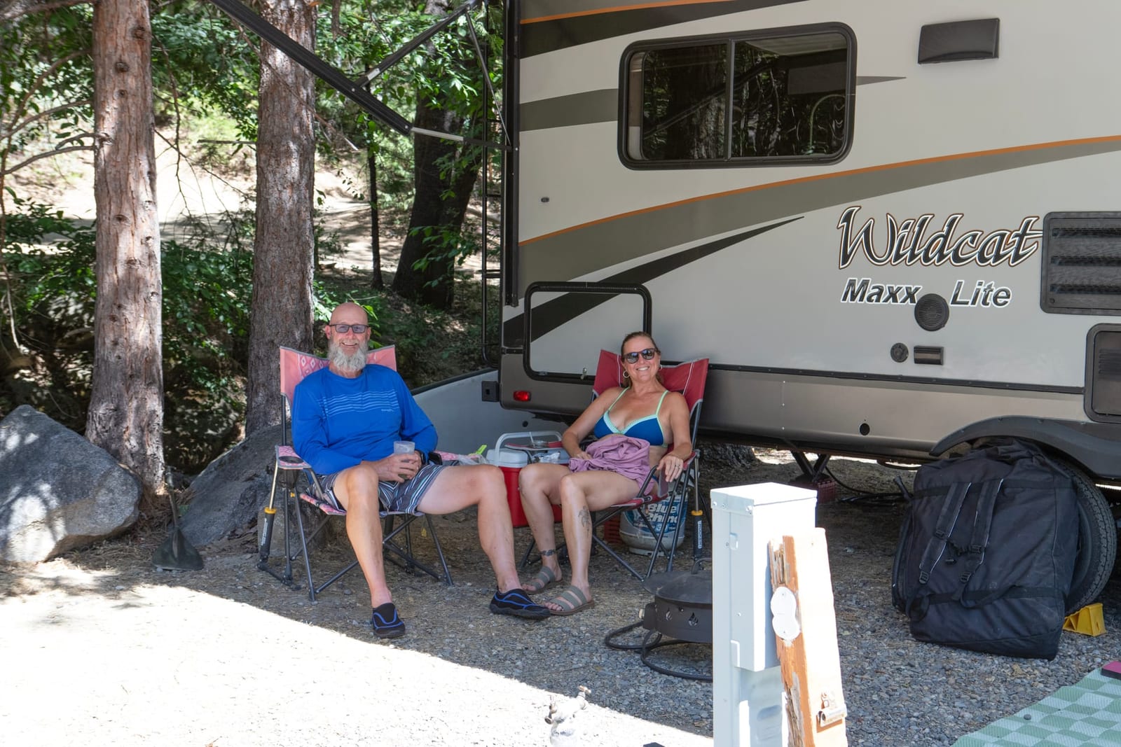 Travel trailer with awning extended and camp chairs set up at a wooded Yosemite campsite — guests relaxing at their fully-equipped rig